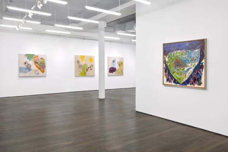 White gallery interior with polished concrete floor, white walls, support columns, and framed artworks displayed at varying heights on walls and columns.
