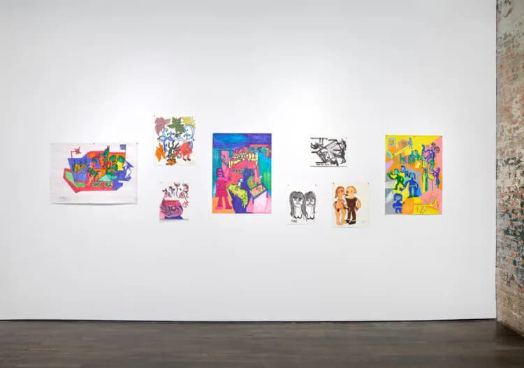 White gallery wall displaying seven small colorful artworks of mixed styles arranged horizontally, including prints with vibrant blues, reds, yellows, and figurative imagery.