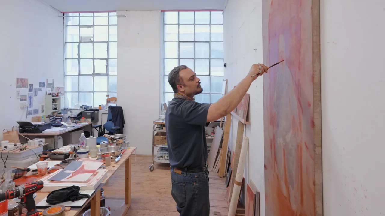 Artist in black shirt painting on a large pink-toned canvas in a bright studio with wooden work tables and gridded windows.