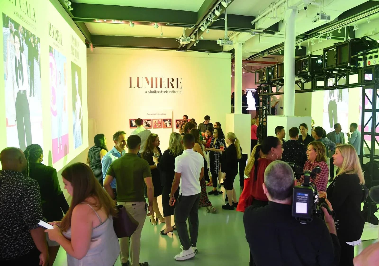 Gallery event with crowd of visitors gathered around a yellow wall with 'LUMIERE' text and illuminated displays in green-lit industrial space.