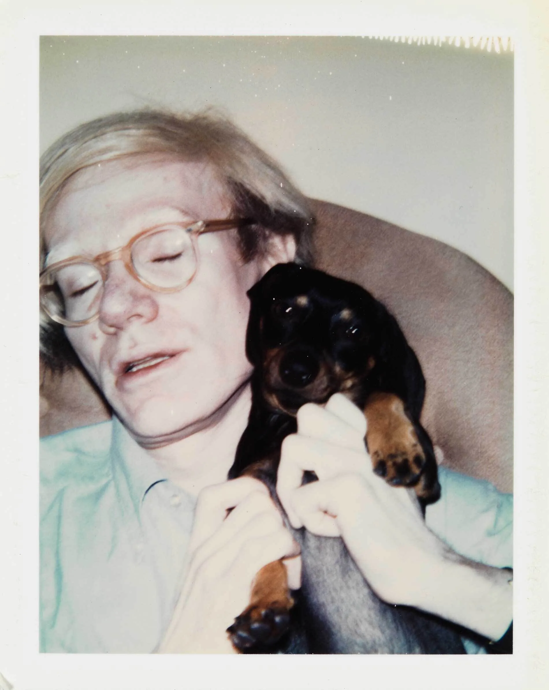 Polaroid-style portrait of a person wearing glasses holding a small black and tan dog, soft color palette.