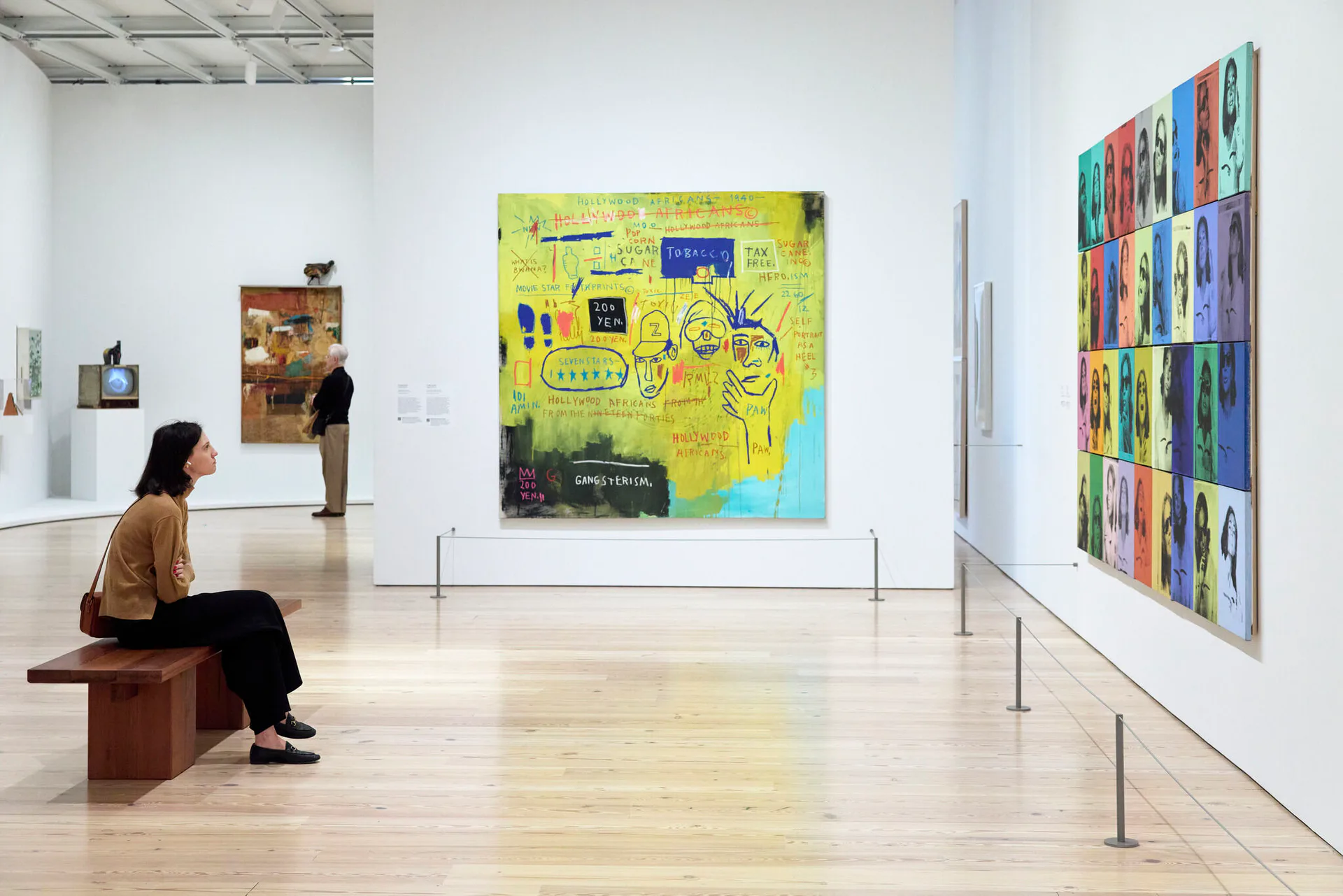 Interior photograph of Whitney Museum gallery showing visitor viewing colorful abstract paintings on white walls, minimalist wooden bench seating.