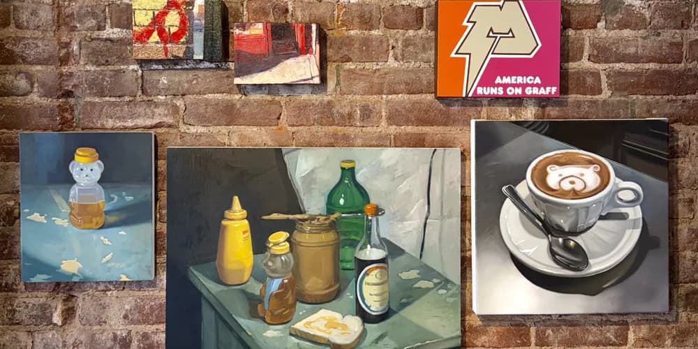 Gallery wall display featuring multiple artworks: still life with bottles and containers, abstract painting, coffee cup photograph, and small colorful pieces with signage.