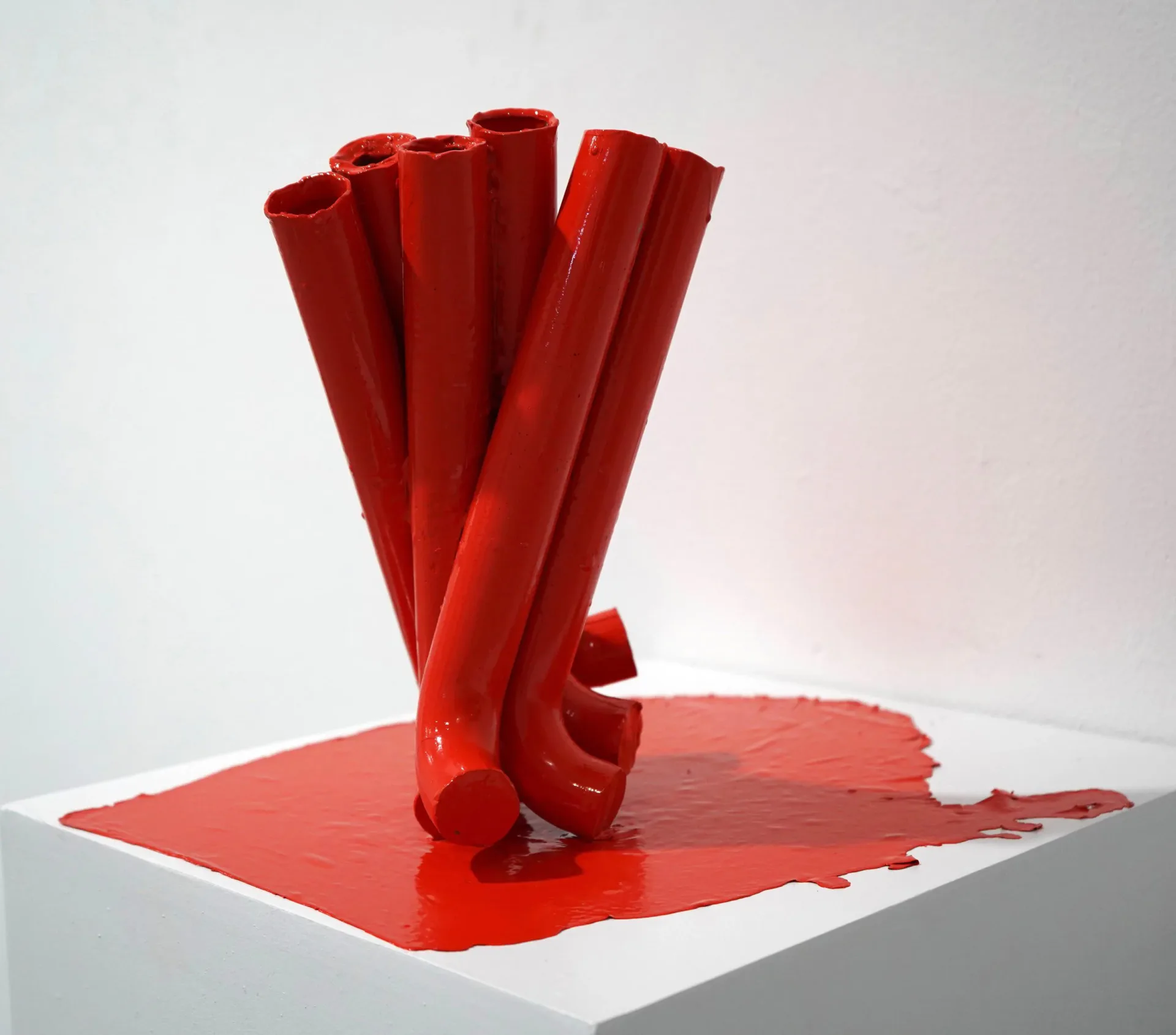 Three-dimensional sculptural installation: red ceramic or resin cylindrical tubes with splayed forms emerging from a red platform, creating an abstract vertical composition.