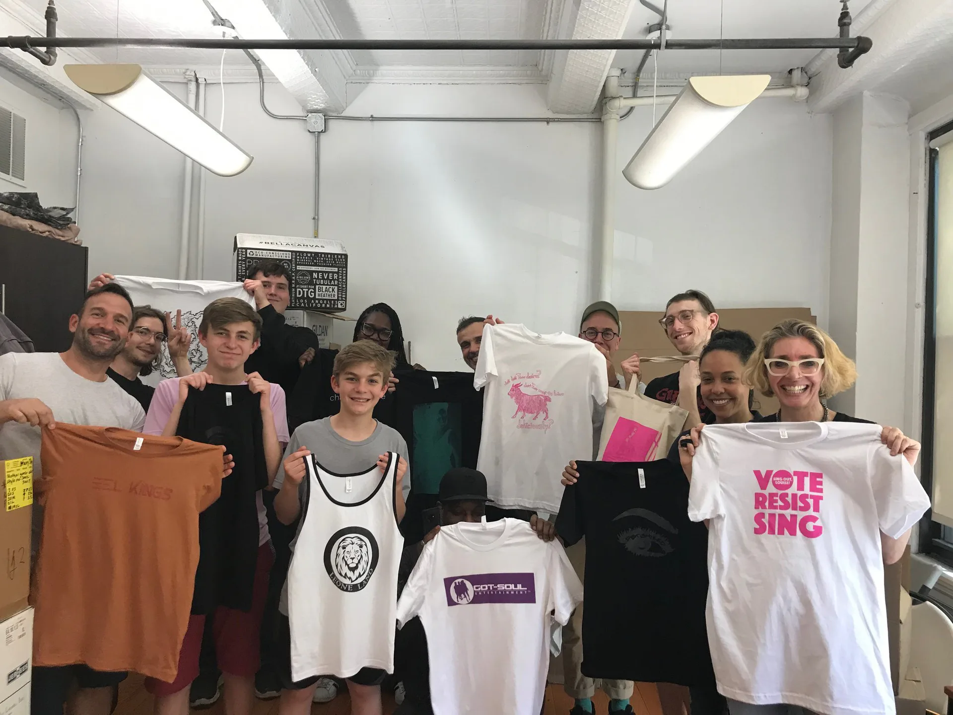 Group of people holding custom t-shirts and merchandise in a bright, fluorescent-lit studio or workshop space at Works In Progress NYC.