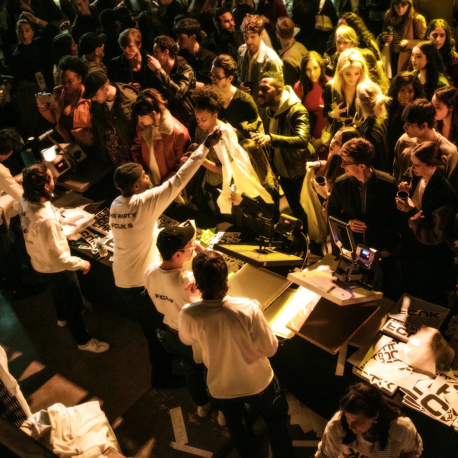 Overhead view of a crowded event or workshop at Works In Progress NYC with participants gathered around tables with printed materials and merchandise.
