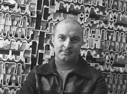 Black-and-white portrait of a bald man with a mustache wearing a dark leather jacket, photographed against a patterned abstract background.