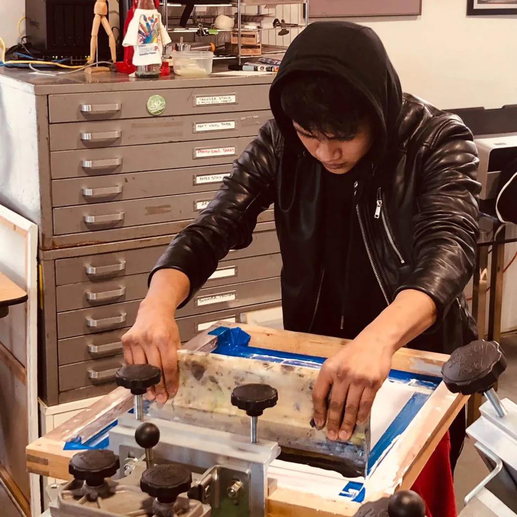 Person in black leather jacket working at a desk with tools and materials in a studio space with metal filing cabinets.