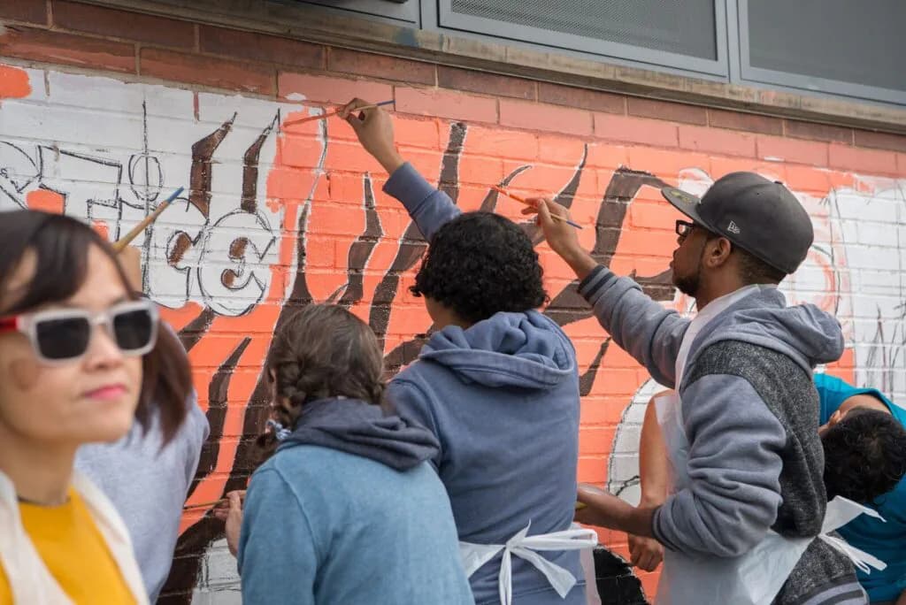 Group of young people painting a mural on a red brick wall outdoors, creating black lettering and designs.